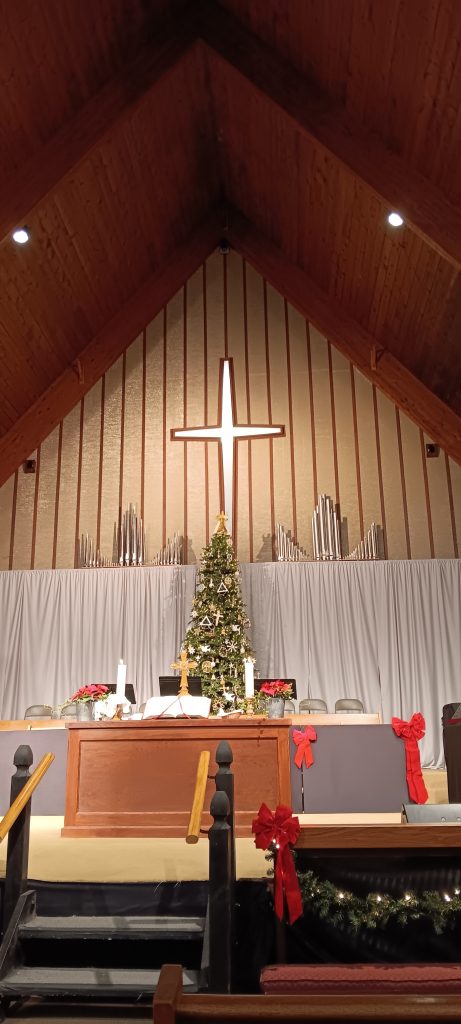 Altar where Michael Robinson worshiped. Christmas tree decorated with white, wooden, and silver ornaments, red bows, a white cross, white candles, and an open book on the wooden table. Steps lead up to the altar and there's a curtain and organ pipes in the background. 