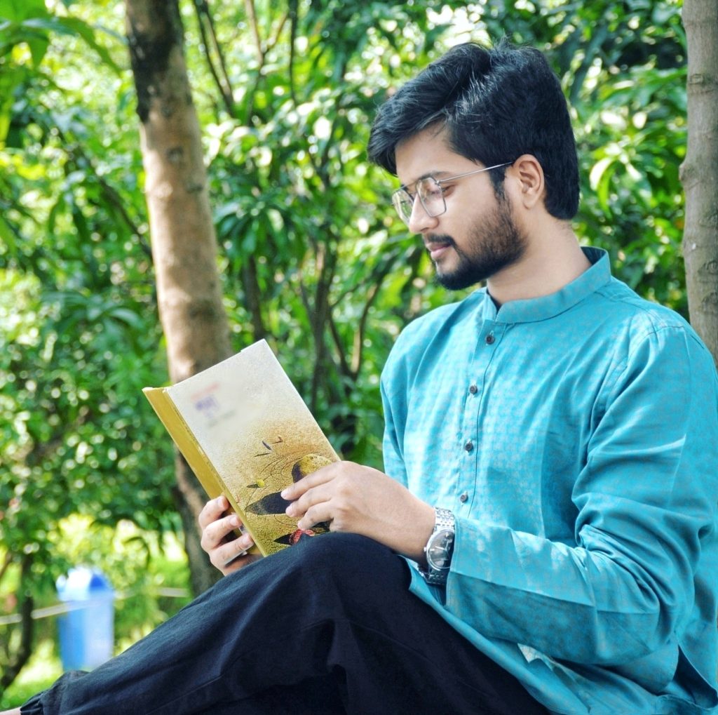 South Asian man with short brown hair, a trimmed mustache, reading glasses, and a blue collared short and dark slacks and a wristwatch sitting outside under green leafy trees reading a book. 