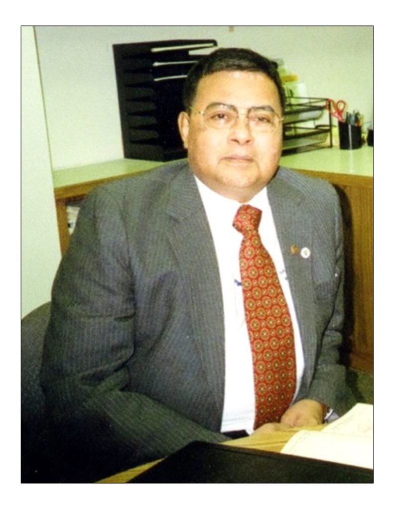 Middle aged South Asian man in a gray suit with a white shirt and a red and orange patterned tie. He's seated at a desk in an office. 