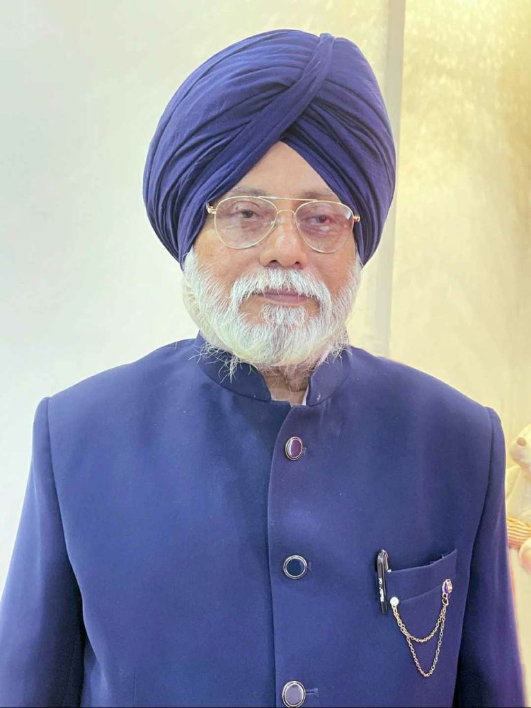 Older South Asian man with a purple turban, a white beard and mustache, reading glasses and a purple suit coat.