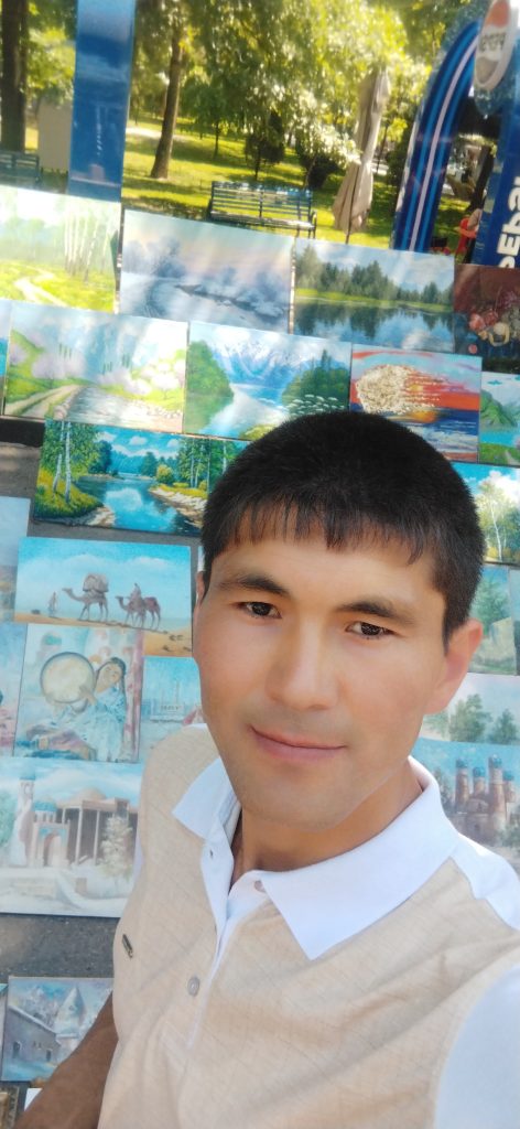 Central Asian teen boy standing in front of a wall of drawings, mostly of nature and waterfalls and Islamic style buildings. He's got short brown hair, brown eyes, and wears a tan sweater and collared white shirt. 