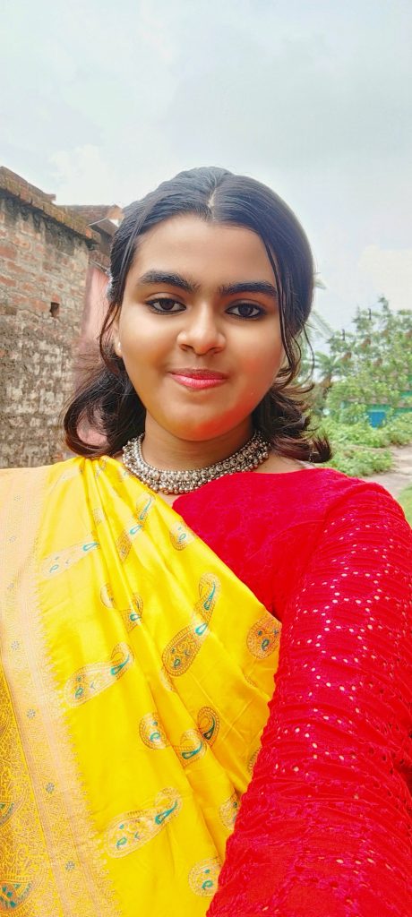 Young South Asian woman with curly dark hair, a necklace, a red and yellow sari. She's standing in front of trees and a brick building. 