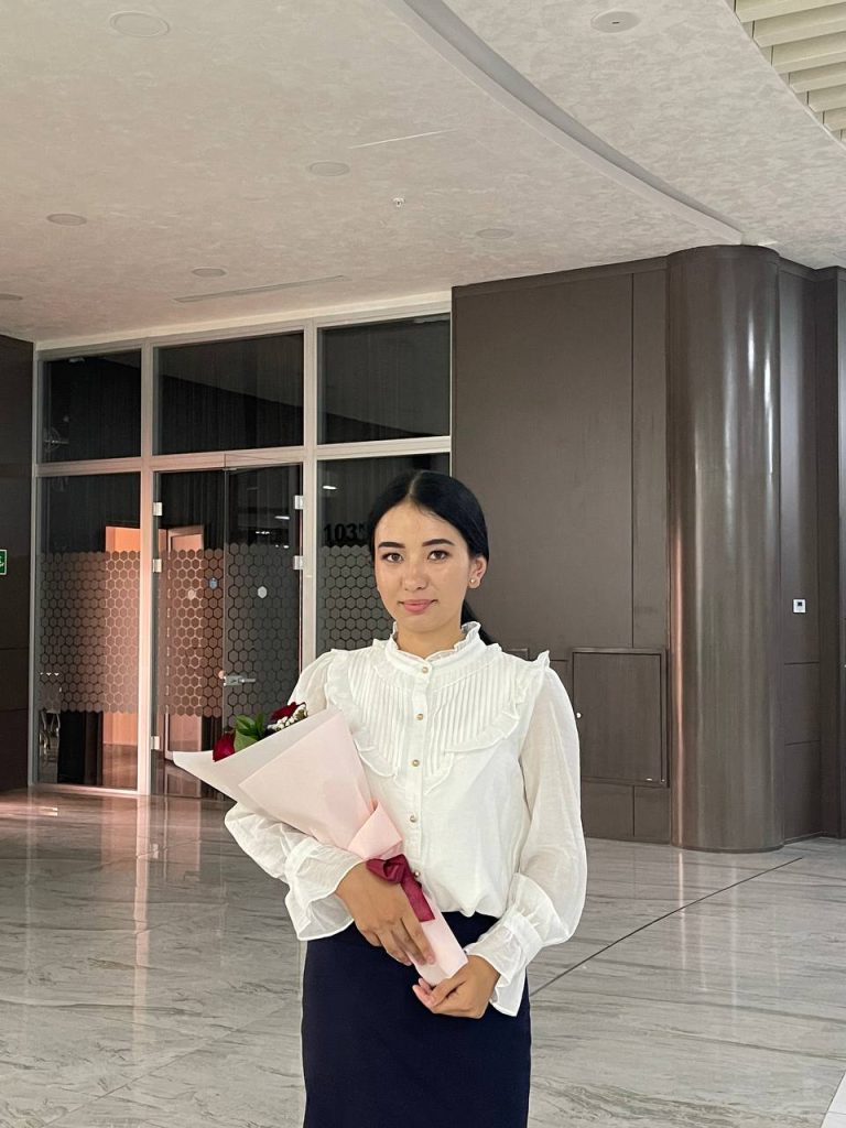 Central Asian young woman stands in front of the entrance to a large modern building. She's got dark hair in a ponytail, a ruffled white blouse and dark skirt, and flowers in her arms.