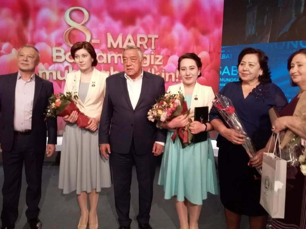 Many middle-aged men and women stand onstage in dresses and suits holding bouquets of flowers. 