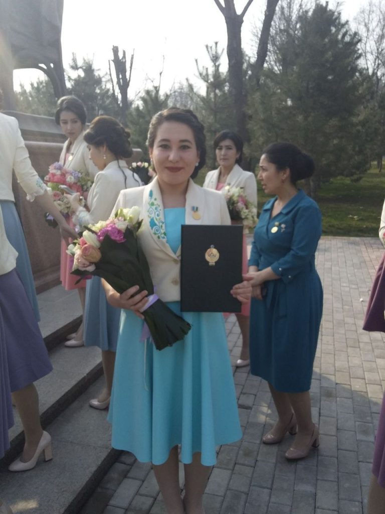 Central Asian woman in the midst of other people. She's in a white coat and dark hair behind her head in a blue dress and a white coat holding a bouquet of flowers and a certificate. She's on concrete bricks near steps. 