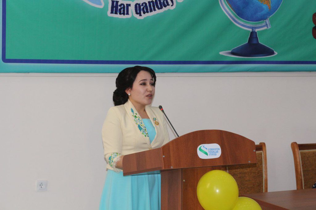 Central Asian woman in a tan sweater with flowers embroidered onto it and a blue dress and dark black hair in a ponytail speaking at a podium with a poster behind her and yellow balloons in front of her. 