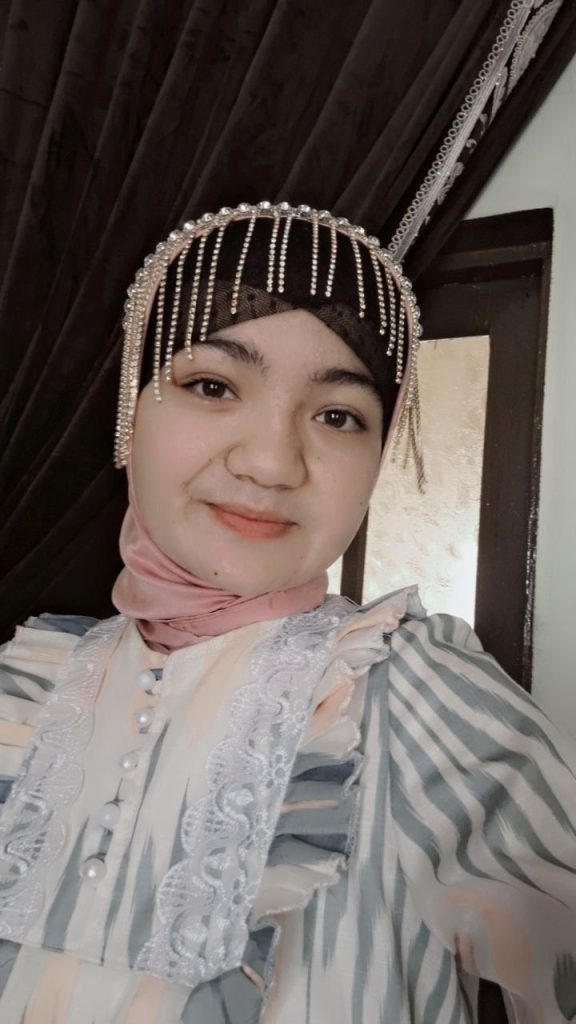 Central Asian woman with a beaded headdress, pink scarf, and ruffled gray and white blouse. 