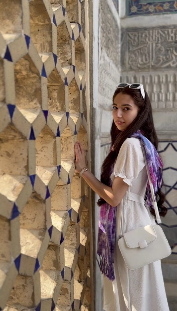 Central Asian teen girl in the corner of a building with stone walls engraved with designs. She's got white sunglasses on her head, long straight brown hair, a white summer dress, a purple tie die scarf, and a white purse and wristwatch.