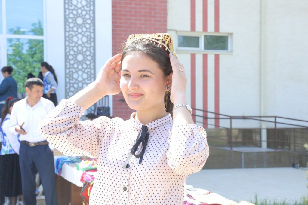 Central Asian young woman with long dark hair and a headdress and a white blouse with black dots and a black ribbon tie. She's in front of a building with others in the background. 