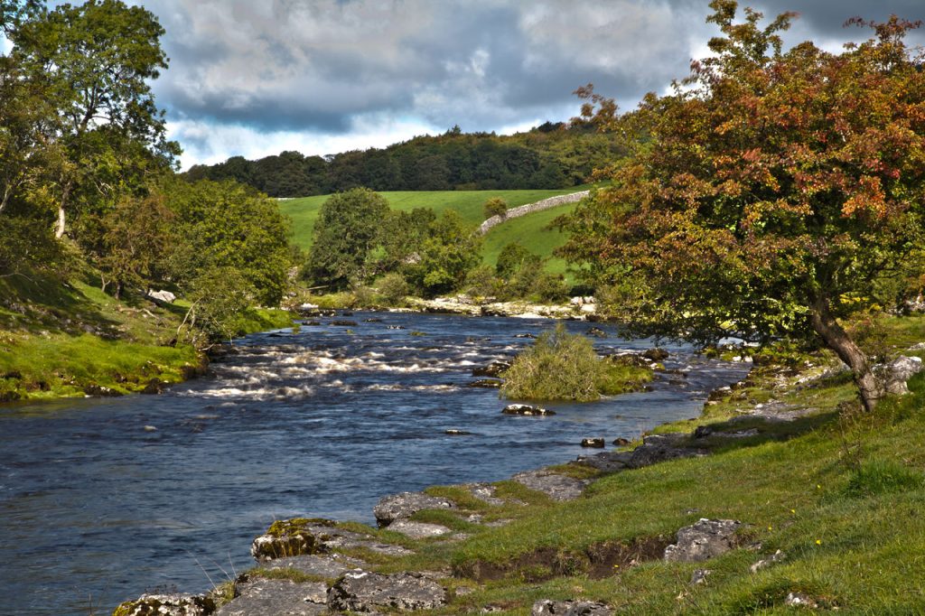 Flowing blue river with rapids over some rocks and grass on either side. Trees and hills in the distance, a few clouds in the sky. Dales of the U.K.