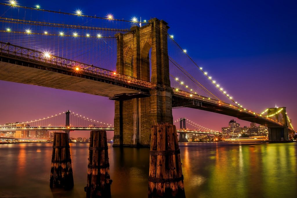 Nearly still river water under the Brooklyn Bridge at dawn or sunset. Sky is blue at the top and pink near the horizon, the bridge is lit up with lights and the city in the distance is lit up as well. 
