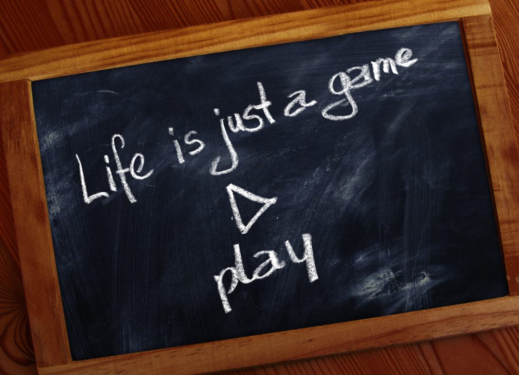 "Life is just a game, play" written in chalk on a blackboard. Blackboard is framed in wood and resting on a wooden table. 