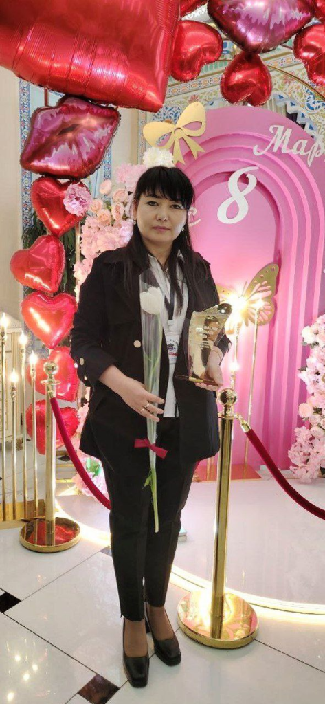 Central Asian woman with long straight dark hair, brown eyes, a black coat and white blouse, holding a white rose and a trophy. She's got balloons and flowers and a pink background behind her. 