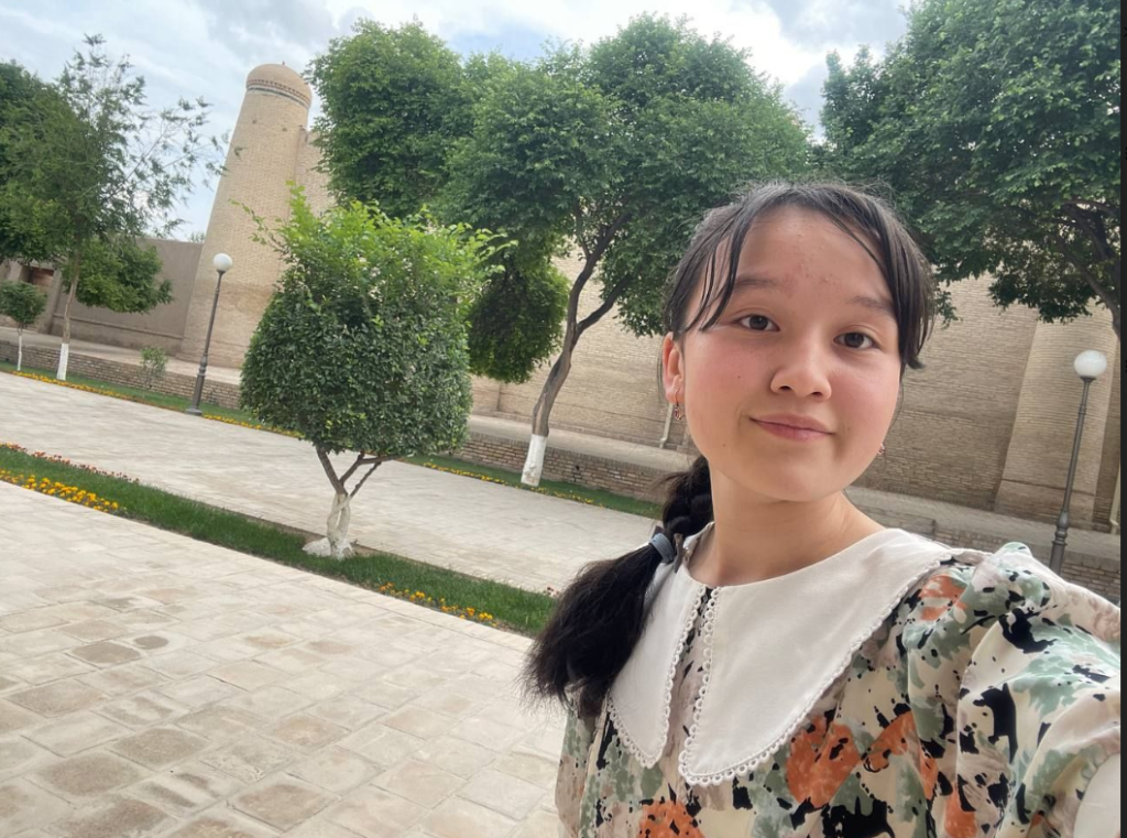 Central Asian teen girl with black hair in a ponytail and a flowered dress in front of a park with a stone building and trees.