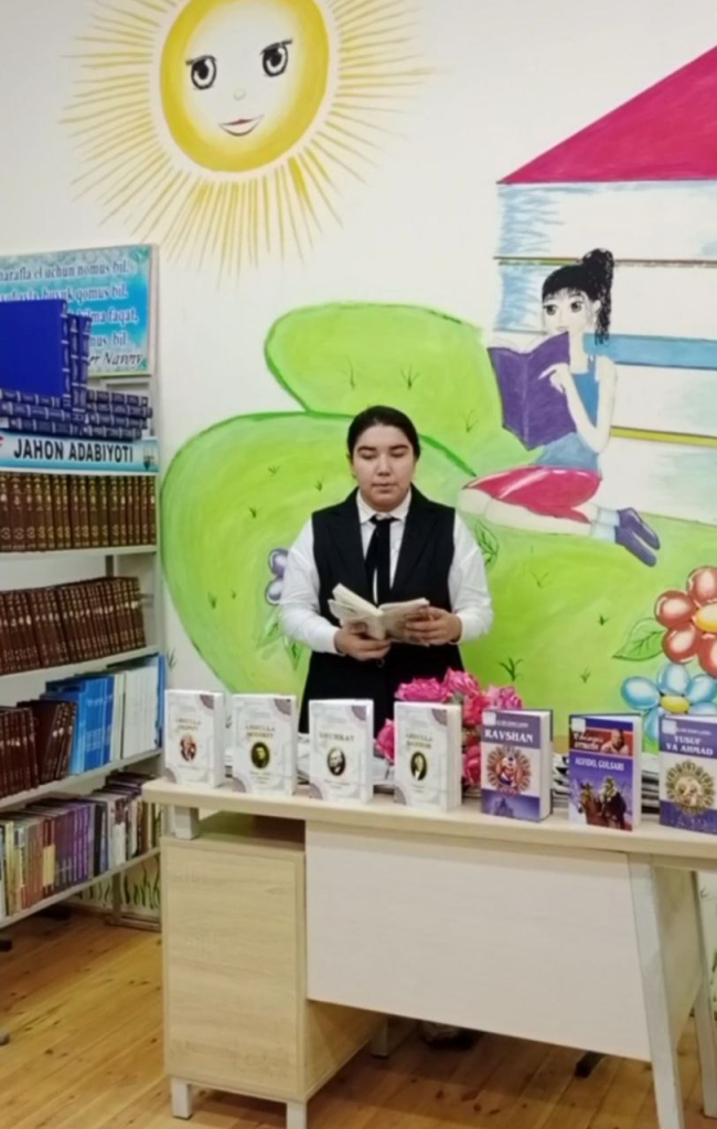 Central Asian woman in a black jumpsuit and tie with a white blouse holding an open book in front of a table with a whole set of books on display. Behind her is a childlike drawing of a girl reading a book on a sunny day on a grassy field with flowers. 