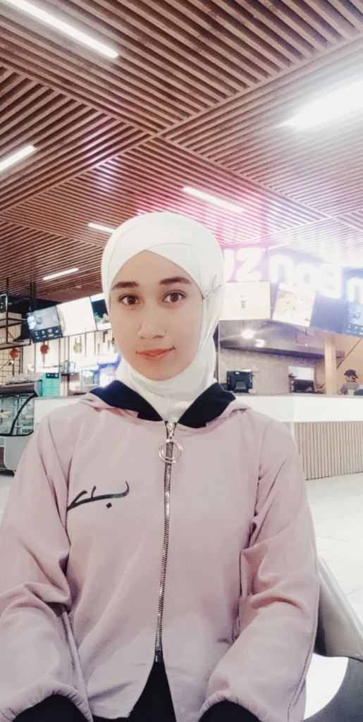 Central Asian teen girl with a white headscarf, pink zipped jacket, and brown eyes standing in front of a set of TV screens. 