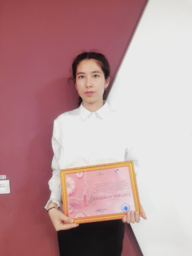 Central Asian teen girl with dark hair up behind her heard and a white dressy collared blouse in front of a pink and white wall holding a framed certificate. 