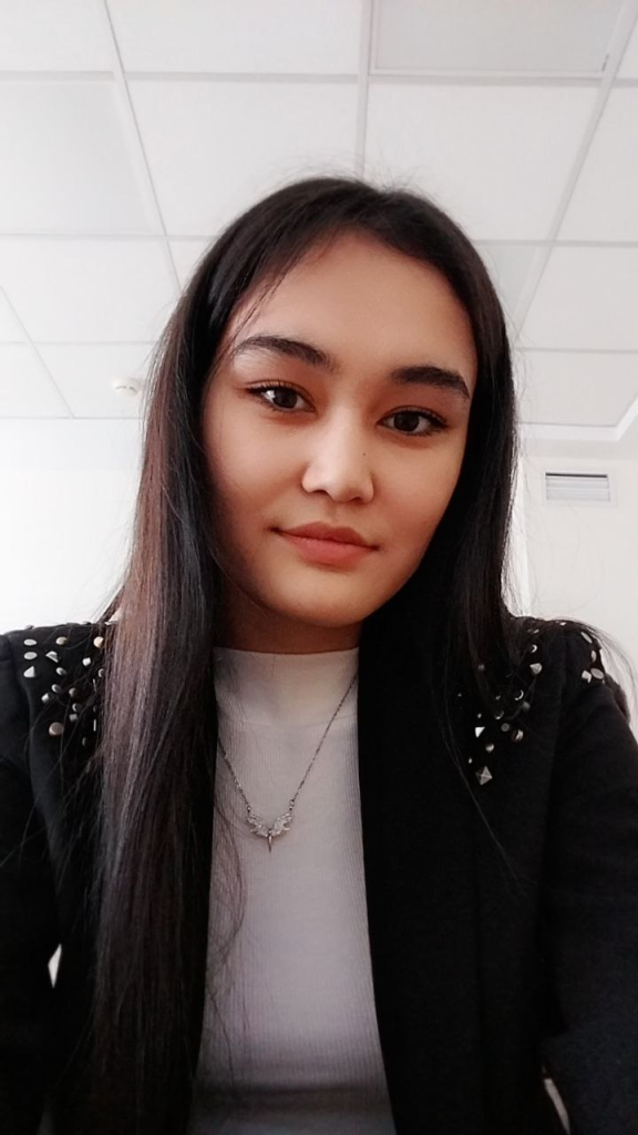 Teen Central Asian girl with long straight dark hair, brown eyes, a white sweater, a small silver bird necklace, and a black coat speckled with silver pieces on the shoulders. 