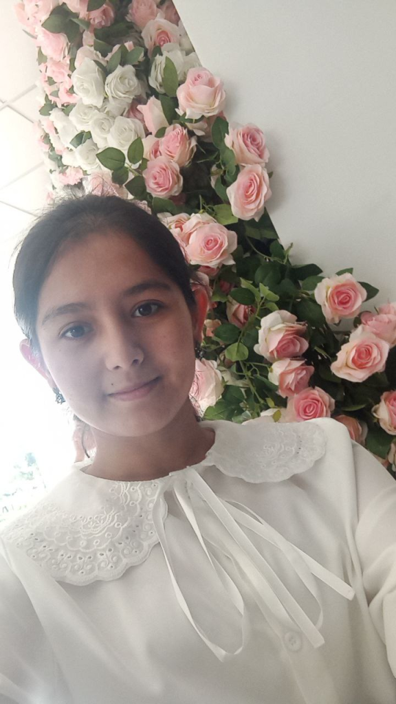 Central Asian teen girl with her dark hair up in a bun and a white lacy ruffly blouse. Pink roses are behind her. 