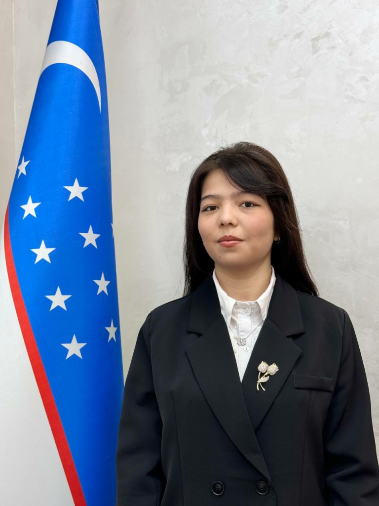 Central Asian woman with long dark hair, a white collared shirt, and a black coat standing next to a red, white, and blue flag. 