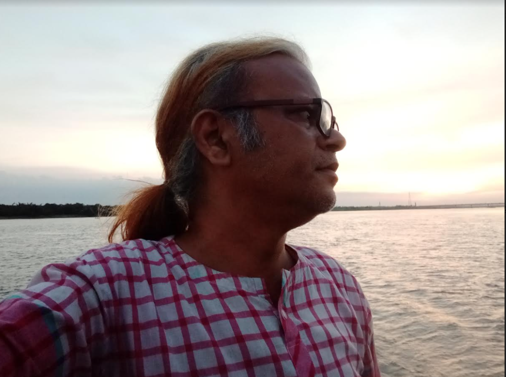 Middle aged South Asian woman in a pink and white checkered shirt, blond highlights in his dark hair, sunglasses, a ponytail, looking off the distance towards the sun on a lake.