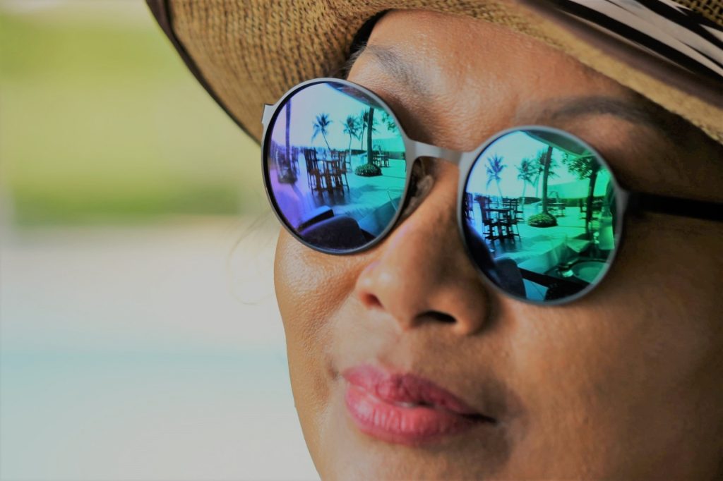 Middle aged Asian woman on vacation with palm trees and tables and chairs reflected in her sunglasses. She's got a straw hat on and the immediate background is fuzzy. 