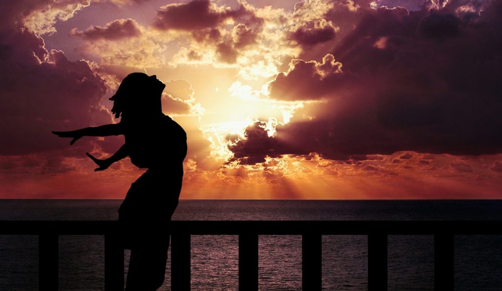 Woman in silhouette stretches her hands back behind her and looks up to the sky on a pier over water at sunrise or sunset. Clouds hide the sun but send out shafts of light. 