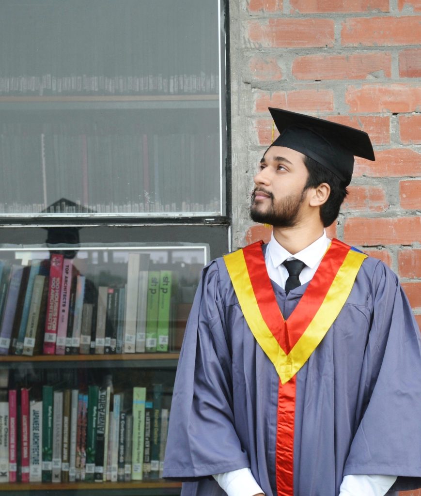 South Asian man, college student age, looking to our left in a graduation cap and gown in front of a brick building and a bookshelf. 