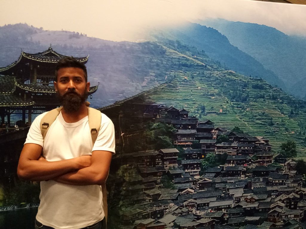 South Asian man in a white tee shirt with a backpack standing in front of a hillside with pagodas and houses built into the grassy terraced hill. 