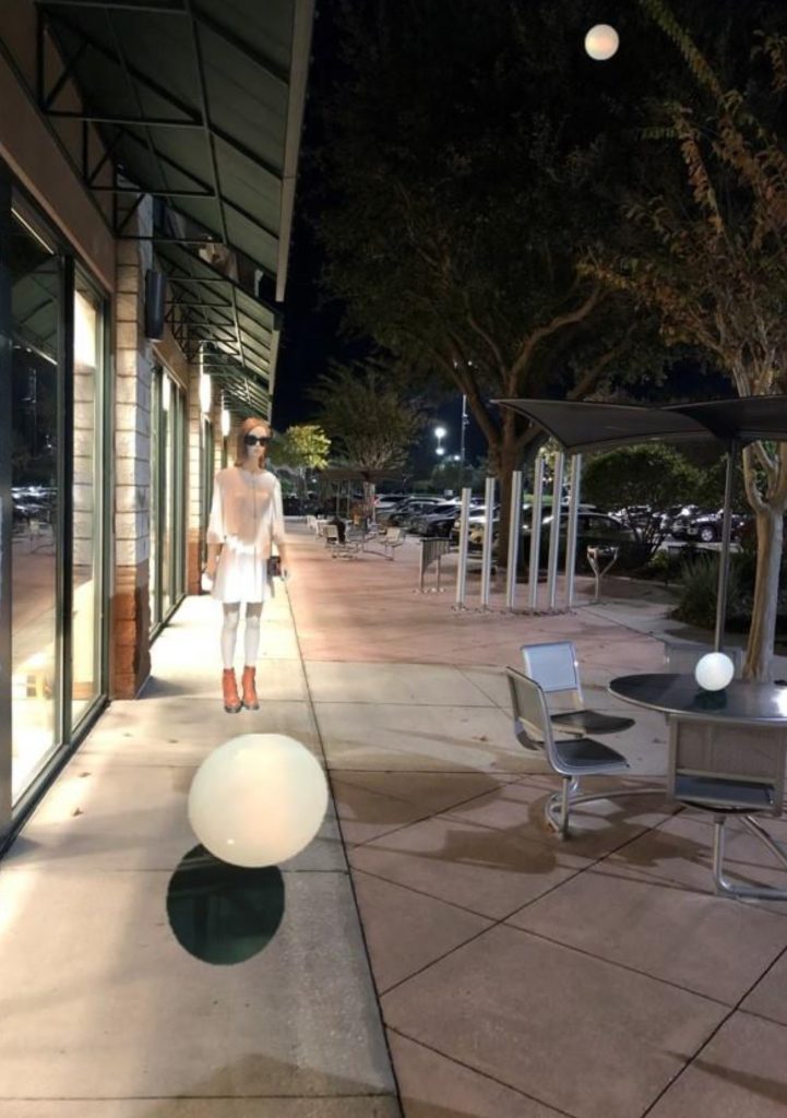 Skinny woman with sunglasses and a white dress and skirt and tan boots under the eaves of a building at night. Tables and trees near her. 