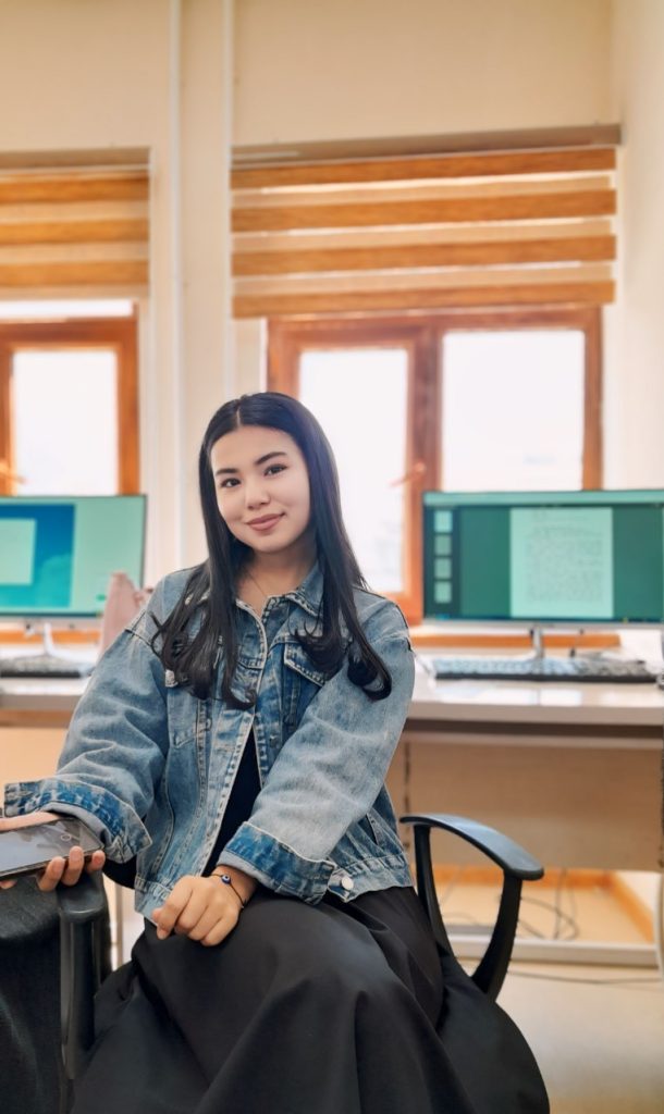 Young Central Asian woman with long dark hair, a jean jacket, and a black skirt in a wooden room with open windows and computers. 