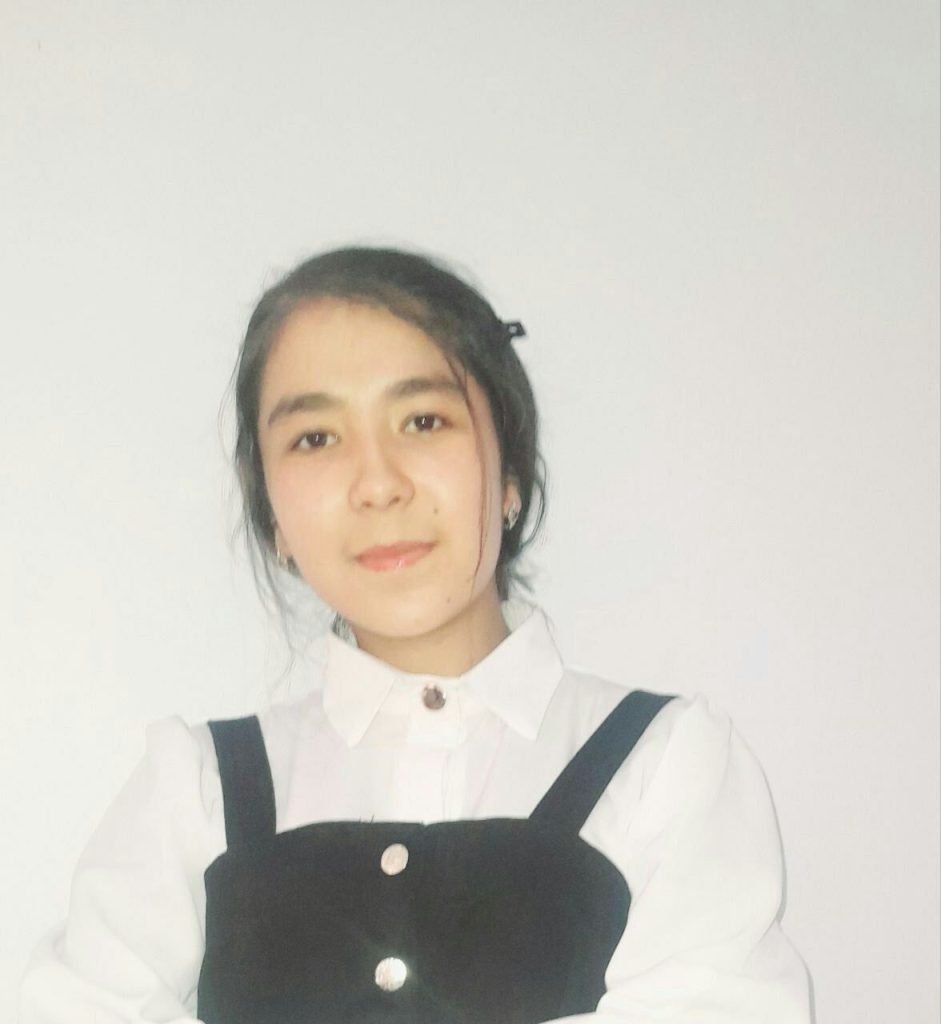 Central Asian girl with dark hair up in a bun and a white collared blouse under black overalls.