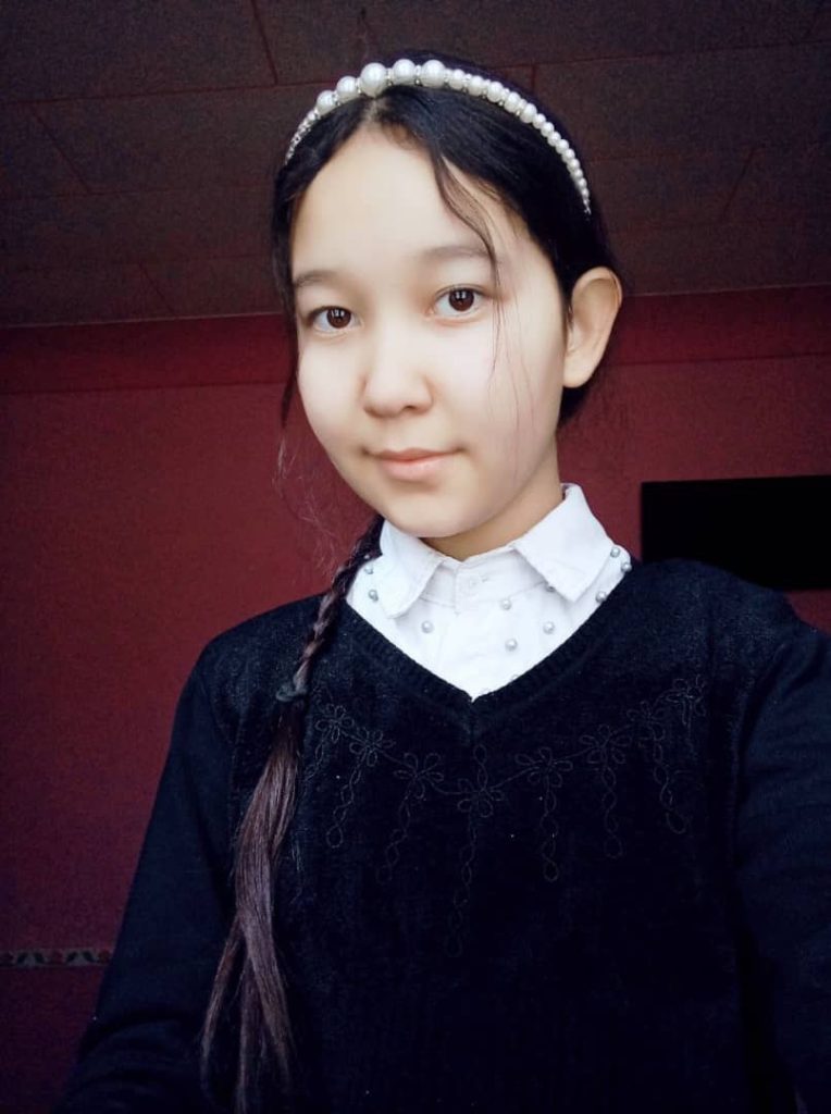 Central Asian teen girl with dark hair and a white beaded headband and a white collared shirt and dark sweater. 