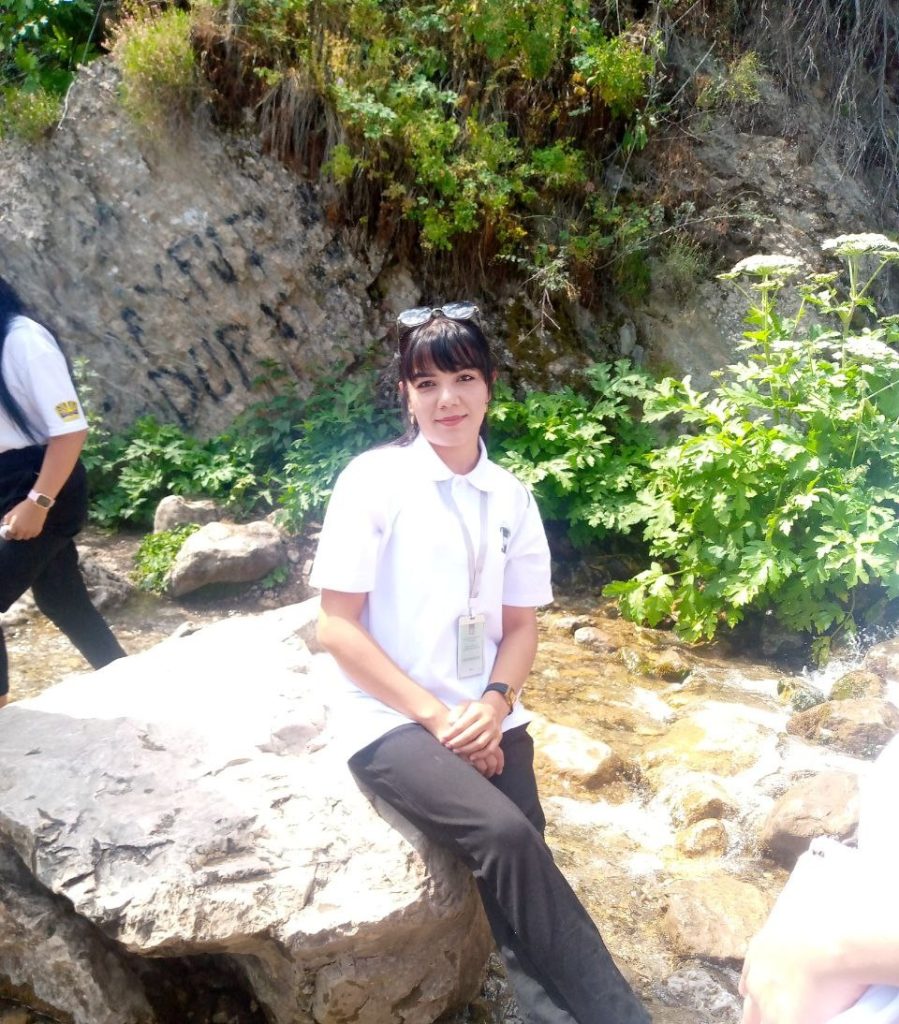 Central Asian woman with short straight dark hair, brown eyes, and a white collared shirt and black pants seated on a rock outdoors with water and green bushes behind her. 