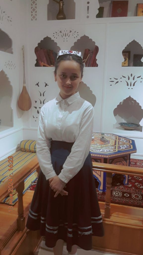 Teen Central Asian girl with a headdress, her hair up in a bun, and a white collared shirt standing in a corner of a room with books in alcoves shaped like Central Asian buildings. There's a carpet and a table. She's in a black skirt. 