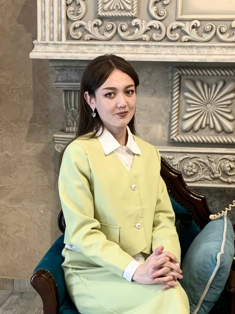 Young Central Asian woman with dark straight hair, small earrings, brown eyes, and a yellow jacket over a white collared shirt. There's a couch and a filigreed column behind her. 