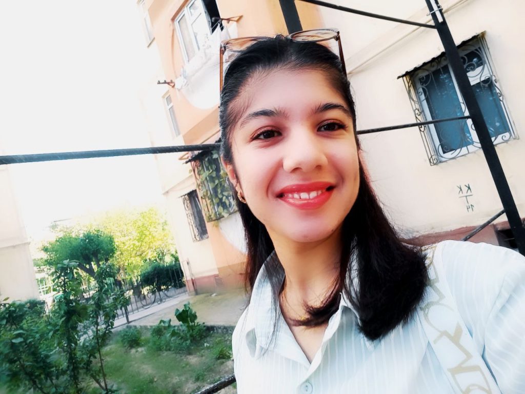 Central Asian teen girl with dark hair and reading glasses and a light collared blouse in front of an apartment building and a garden patch. 