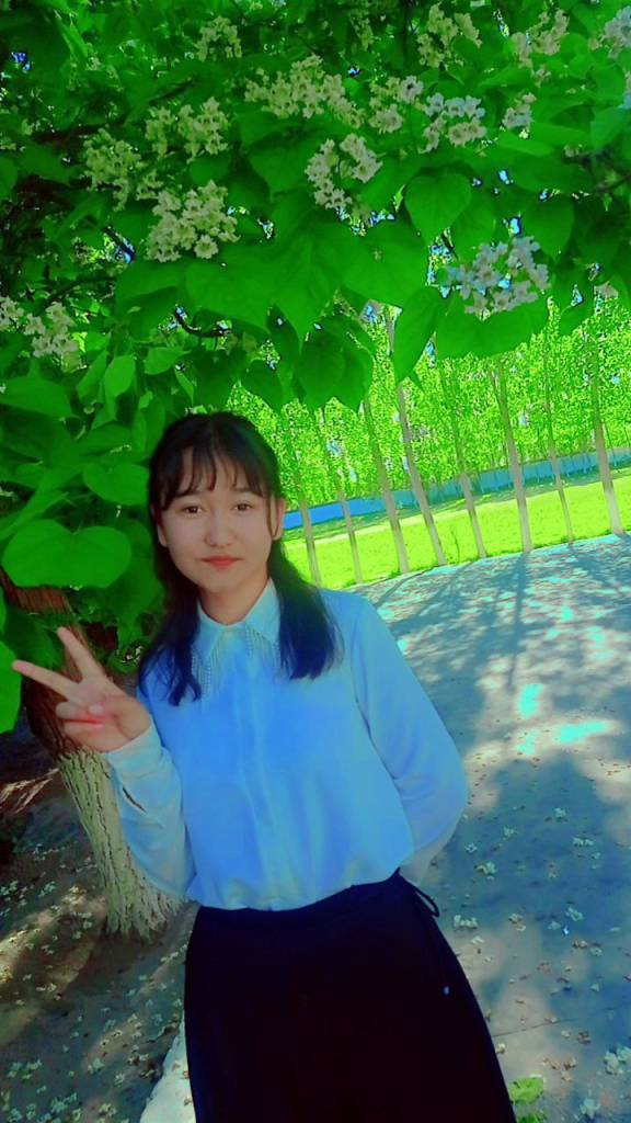 Teen Central Asian girl under a leafy tree with white flowers. She's got long dark hair and a white collared blouse. 