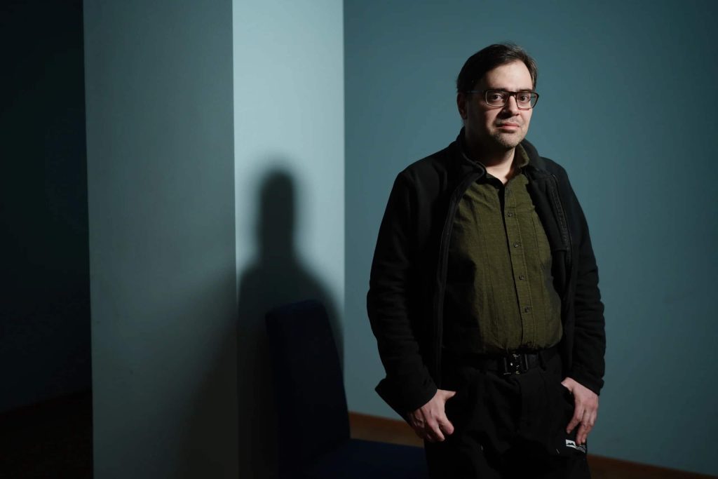 White man with reading glasses, short brown hair, a black coat, slacks and a belt, and a green collared shirt with buttons, standing inside with his shadow against a white wall. 