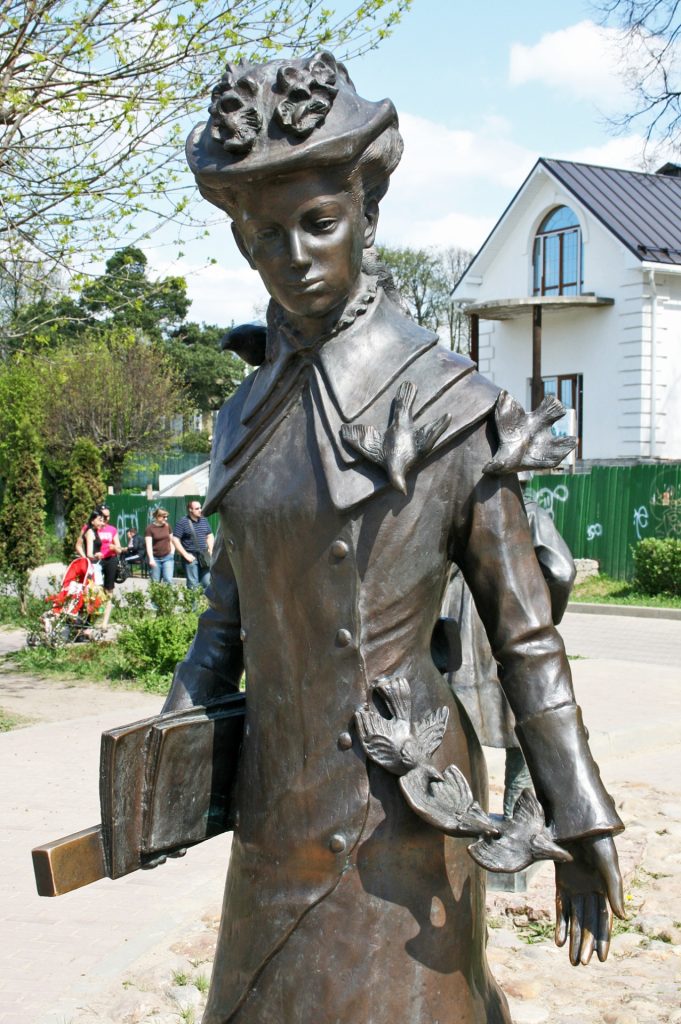 Monument in Moscow, metal statue of a woman with short hair and a feathered hat, with birds landing on her coat. She's holding papers and a rules and is in front of a small building with trees and people in the background. 