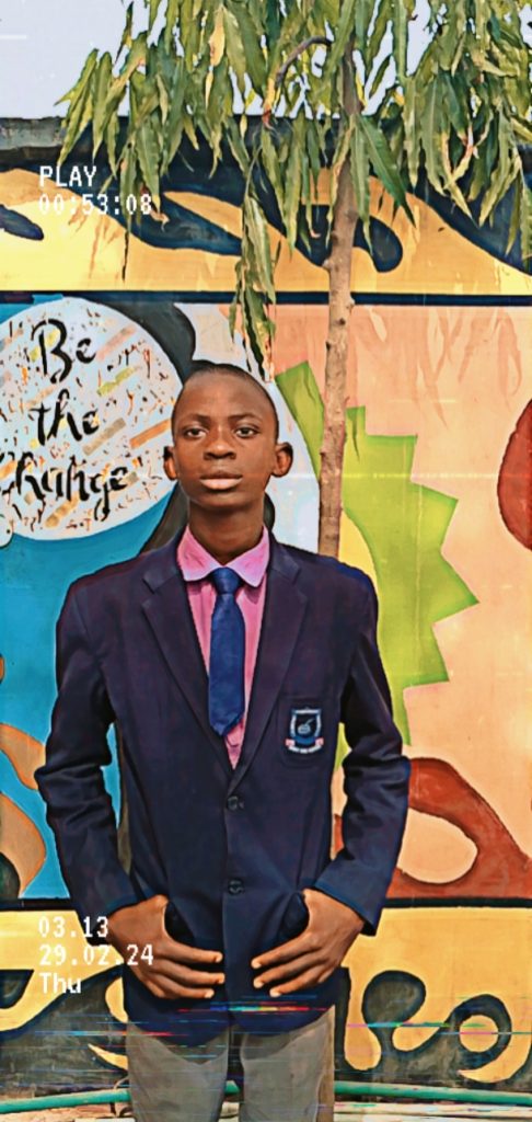 Young Black man in a dark suit, pink shirt, and blue tie in front of a background with a tree that says "Be the Change." 