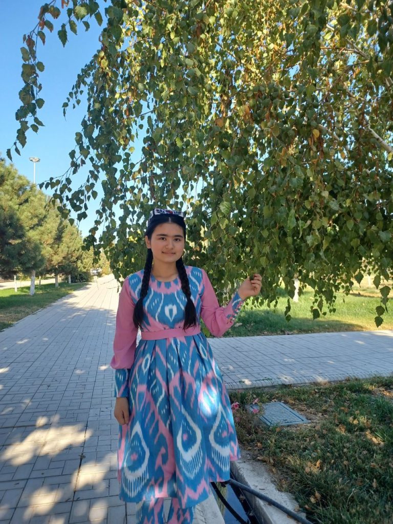 Leafy green tree and concrete path and a Central Asian teen girl in a floral pink and blue and white patterned dress with a black backpack. 