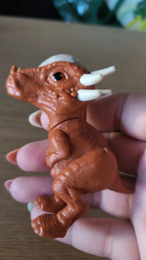 Light skinned person's hand with long fingernails holding a brown tiny toy dinosaur with white horns and a snout.