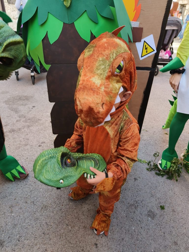 Small child dressed as a brown dinosaur in front of a cardboard and construction paper tree. 