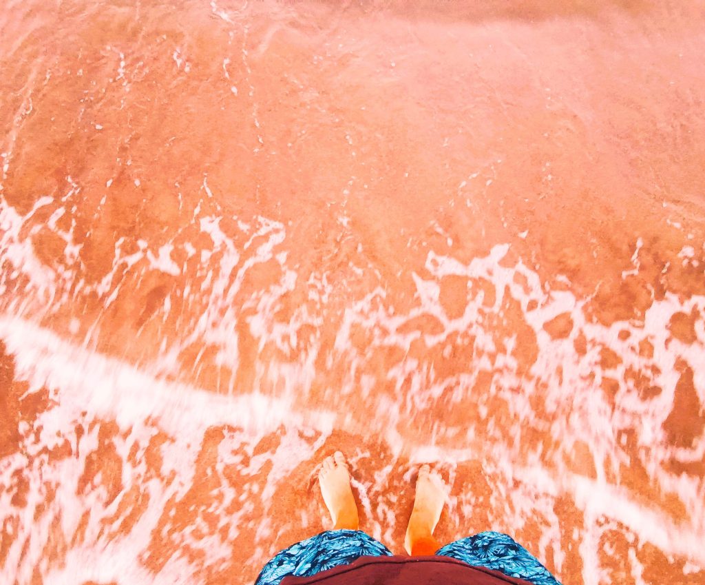 Person's bare feet standing on the beach where the water meets the sand. Orange-red tide, and the person has blue floral-patterned swim shorts. 