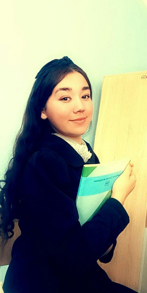 Central Asian teen girl with long dark hair and brown eyes. She's holding a book and is wearing a black coat over a lacy white blouse. 