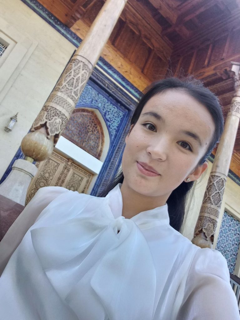 Central Asian teen girl with dark straight hair and a white blouse standing at a diagonal (selfie) in a wood building carved with Islamic style designs. 