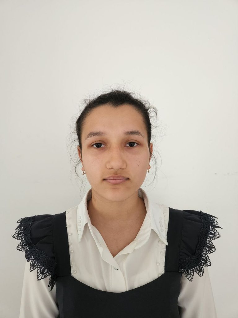 Young Central Asian woman with dark hair in a bun, brown eyes, small earrings, and a white collared shirt and black ruffled vest. 