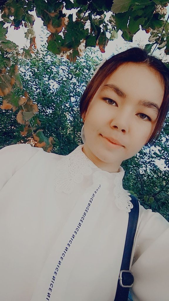 Teen Central Asian girl with short dark hair and brown eyes leaning to the right under a canopy of leafy trees. She's got a lacy white blouse and a black purse.