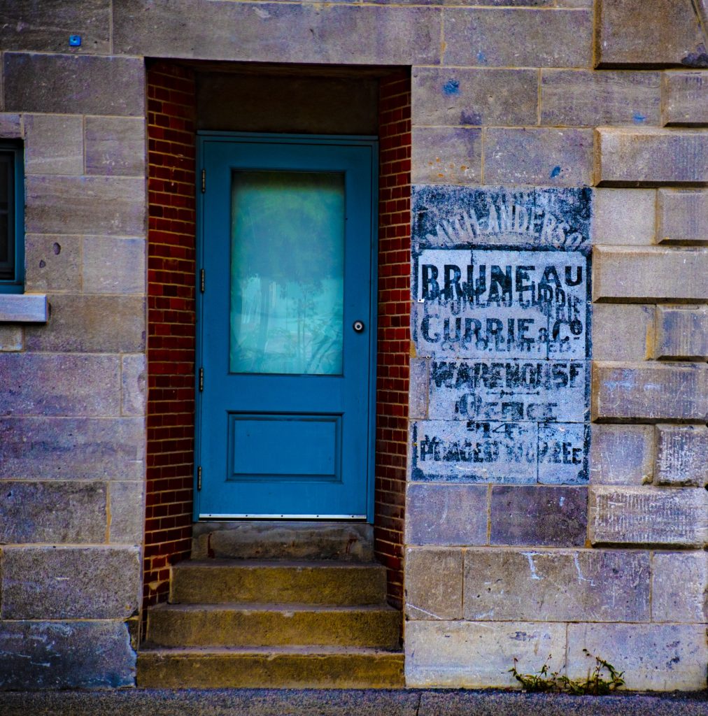 Blue door up a few steps in a concrete brick building with an old faded sign in blue ink on the right of the inset door. 